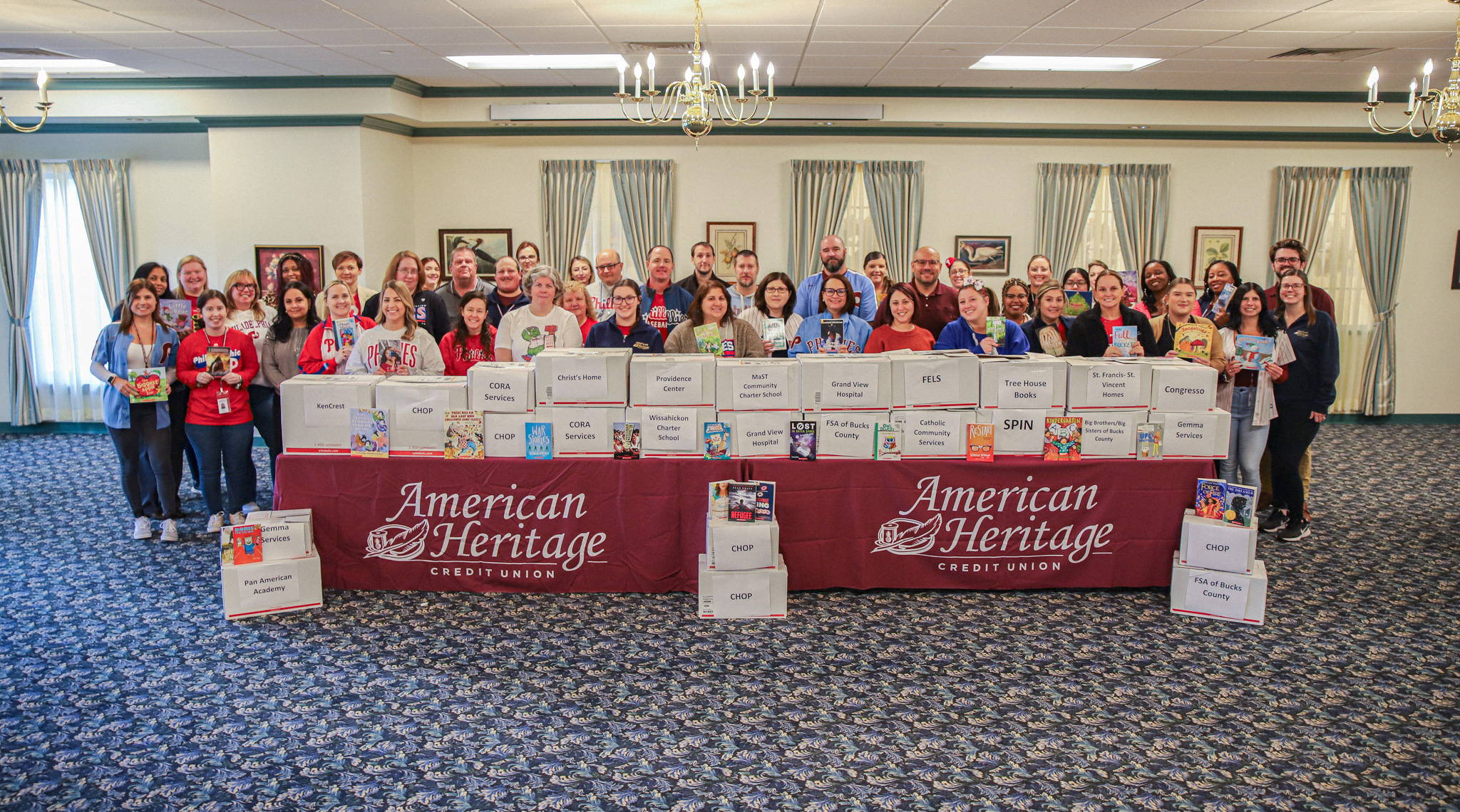 American Heritage’s Books for Kids Committee stand around a table of boxes filled with books to be donated.