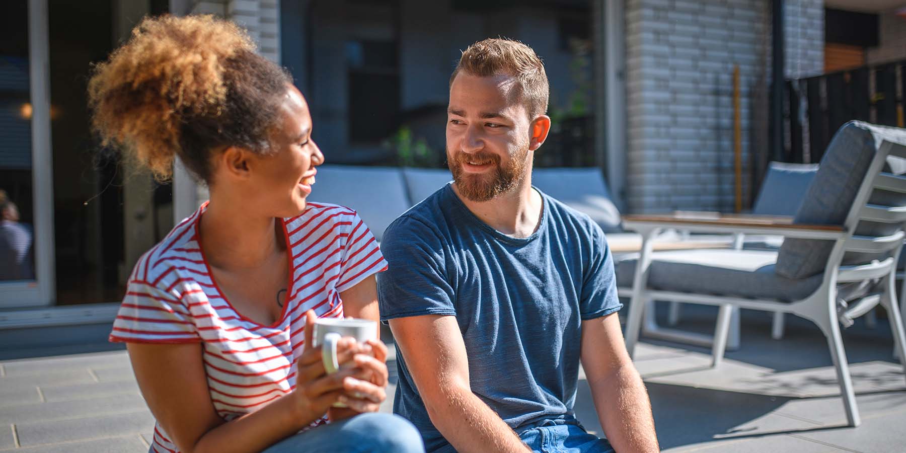 A young couple sitting outside their home, smiling and drinking coffee