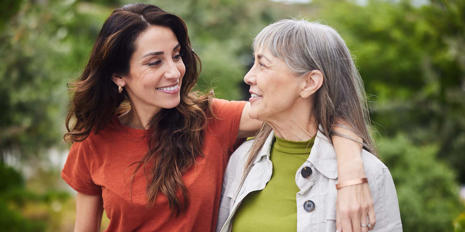 An adult daughter with her arm around her mother, the two smiling at each other