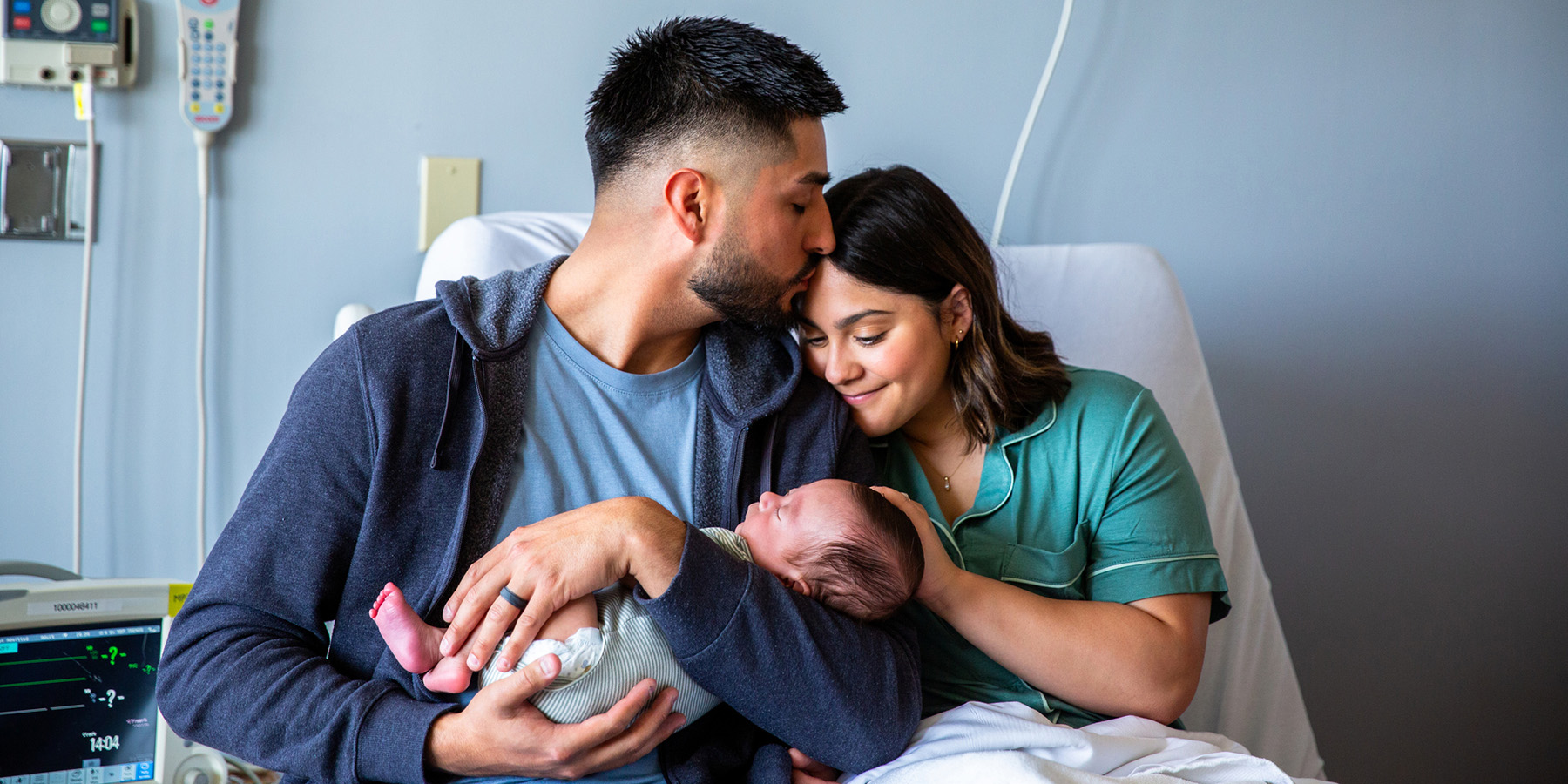 A father kisses his partner as they hold their newborn baby in the hospital delivery room