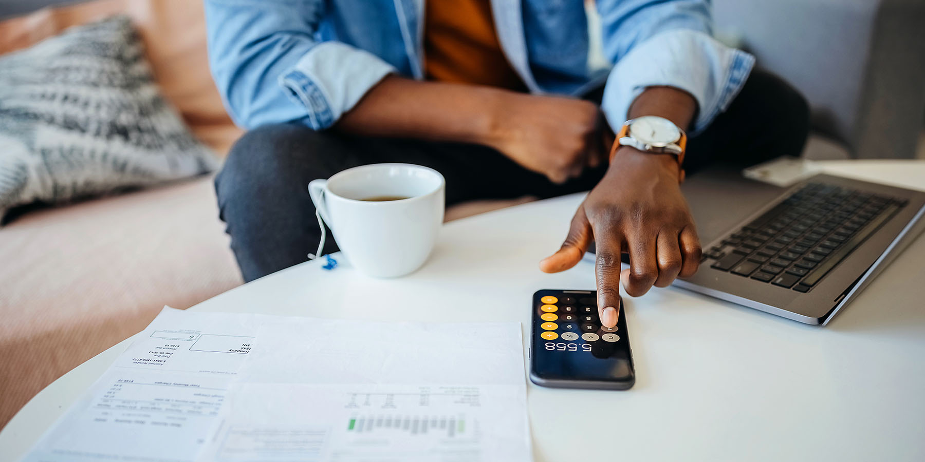 A hand uses the calculator app of a mobile phone, next to an open laptop and cup of coffee