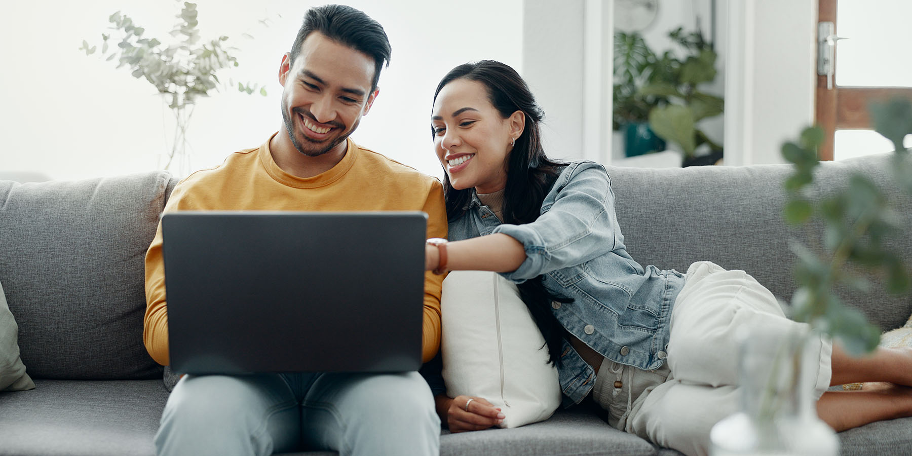 A young, smiling couple sit on their sofa with a laptop, with the woman pointing to the screen