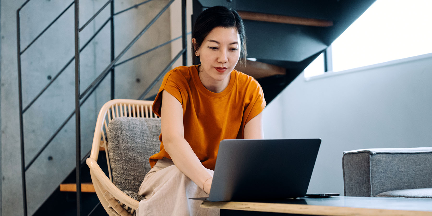 A woman sits in a chair and leans forward, working on a laptop resting on the table in front of her