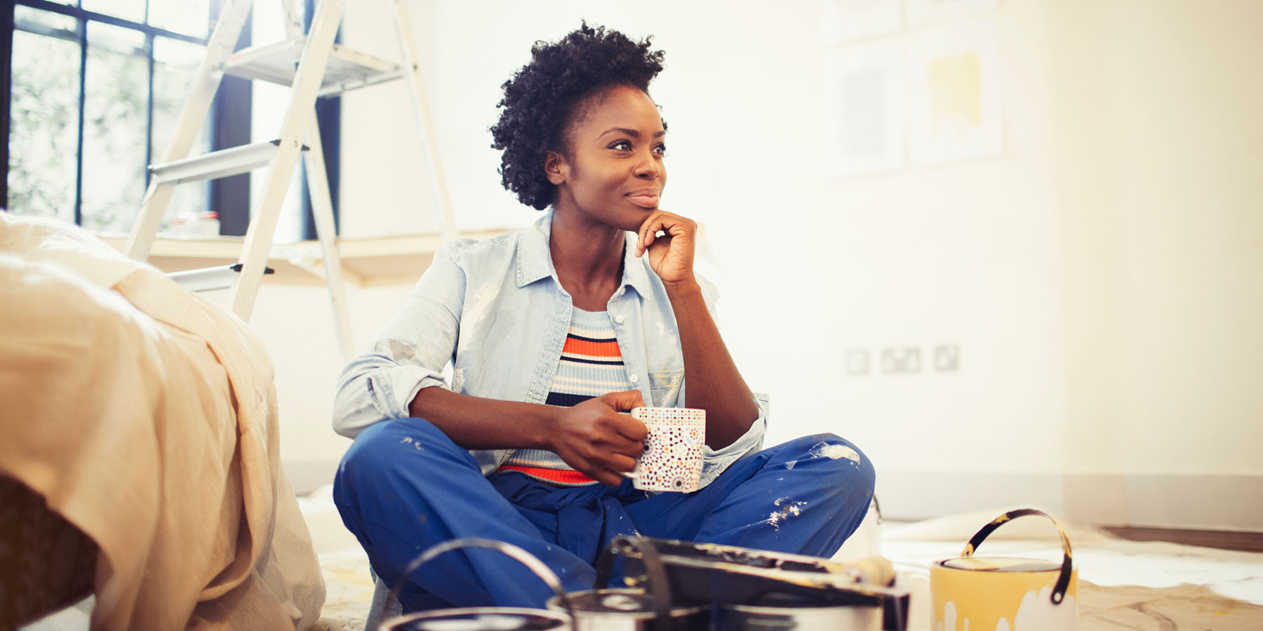 A woman sits on the floor of her home amongst paint supplies, looking thoughtful