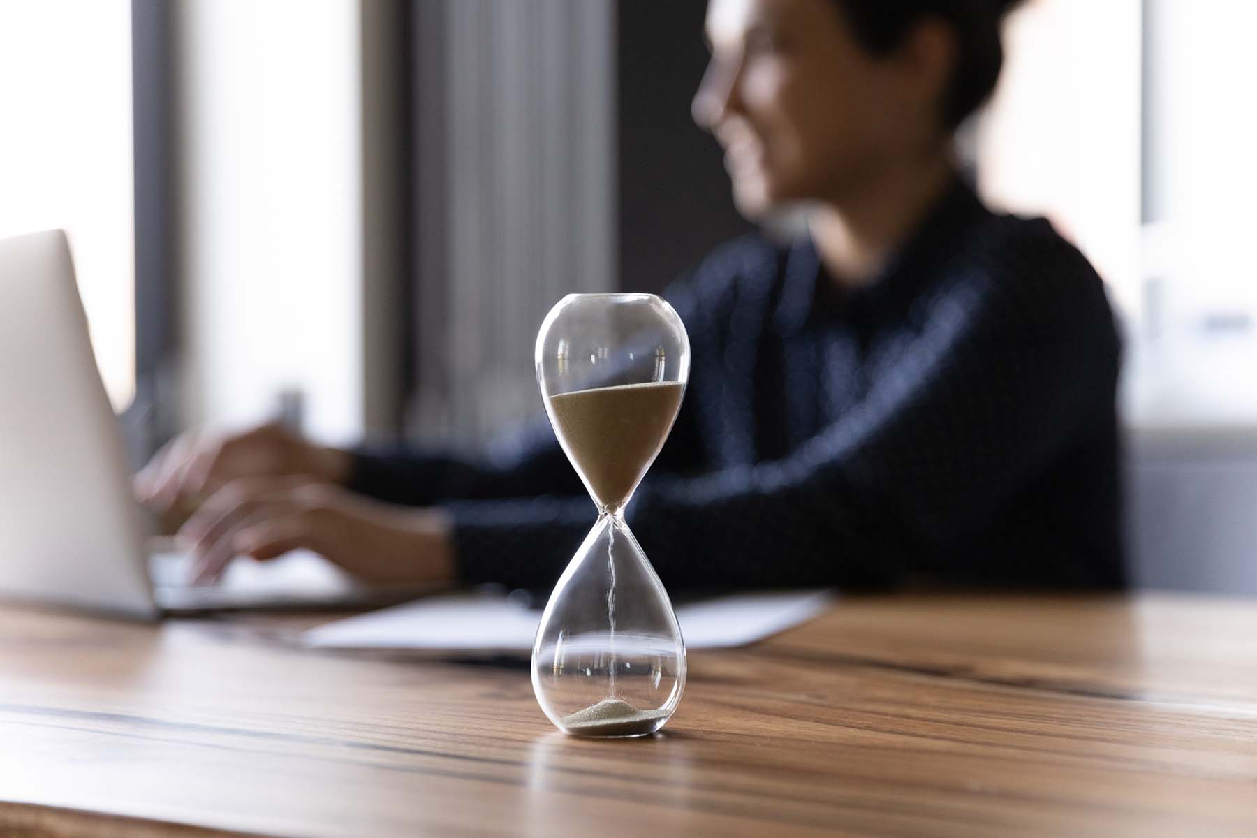 Close up of an hourglass on a desk with a woman using a laptop in the background