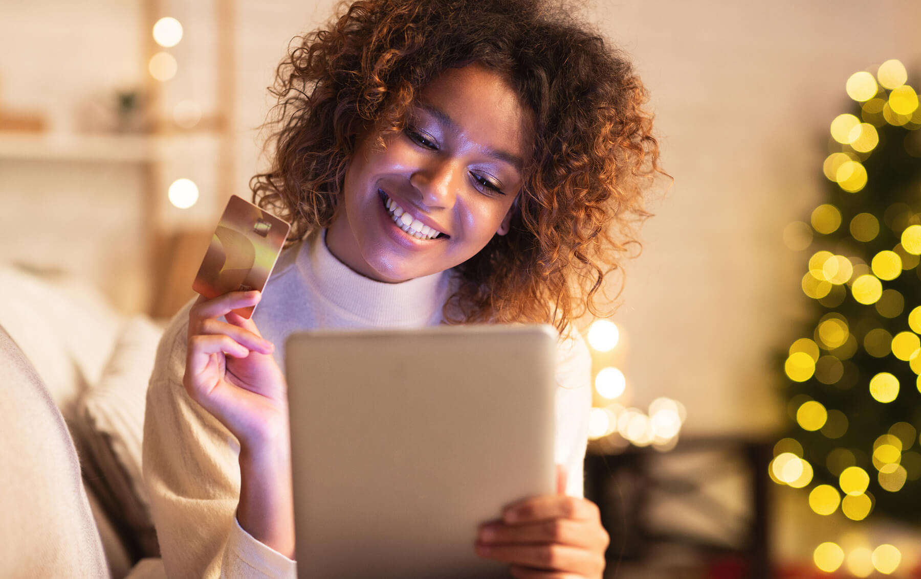 A woman holding a credit card and a tablet beside a lit Christmas tree