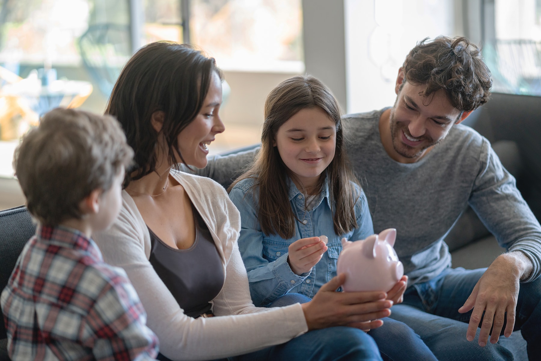 A family of four sits on the couch holding a piggy bank