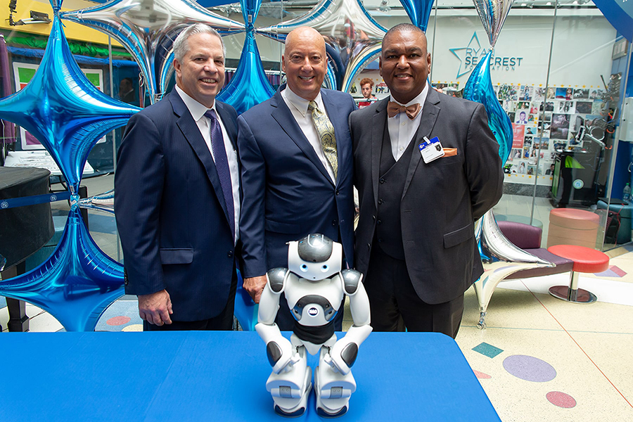 Three men including CEO and President of American Heritage Bruce K. Foulke standing in front of Interactive Robot on table at CHOP